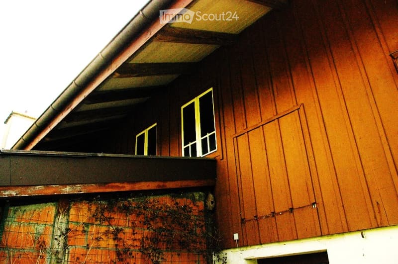 Wooden building exterior with a balcony and windows, showing signs of wear and moss growth on lower sections.
