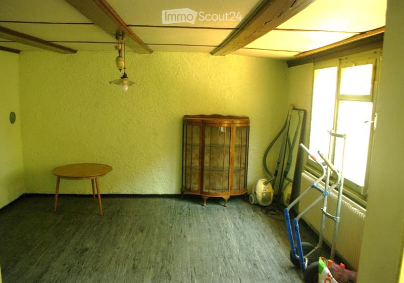 A small, unfurnished room with light green walls and a wooden floor. It features a small round wooden table, a wooden cabinet with glass doors, a vacuum cleaner, a radiator, and some cleaning tools near the window.