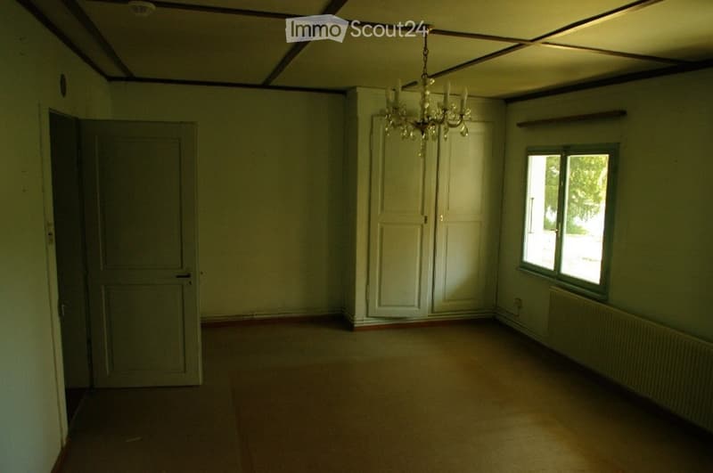 Empty room with wooden flooring, white walls, a single window, a door, and a chandelier hanging from the ceiling.