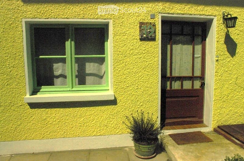 Bright yellow building exterior with a green-framed window and a wooden front door. A small potted plant sits near the entrance.
