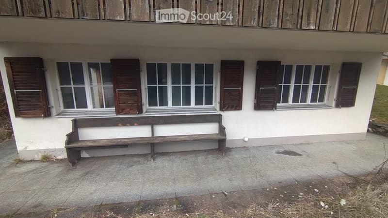 A two-story building with white walls, multiple windows with wooden shutters, and a wooden bench placed near the entrance.