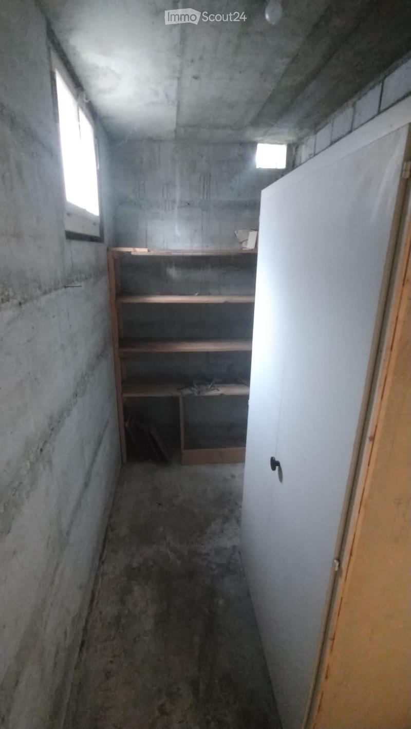 Empty room with concrete walls and floor, small window, wooden shelf, and a partially open door.