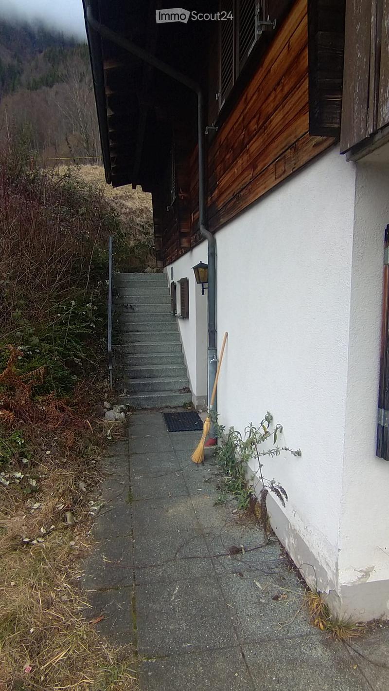 A narrow concrete pathway leading to a raised entrance of a white building with wooden shutters, featuring a small staircase and a broom leaning against the wall.
