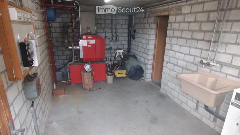 Utility room with exposed brick walls, concrete floor, and various equipment including a red furnace, a sink with a faucet, a wooden block, and tools.