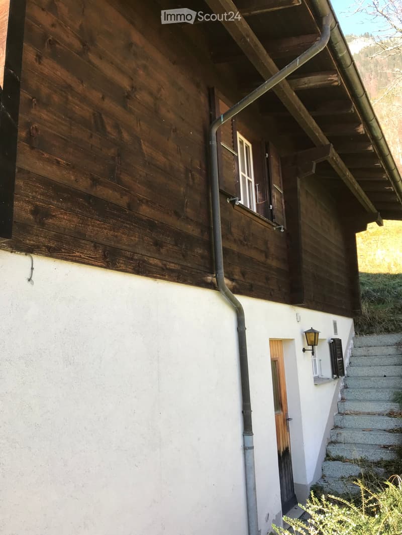Two-story house with wooden siding on the upper level and white plaster on the lower level, featuring a small staircase leading to the entrance and a window with shutters.
