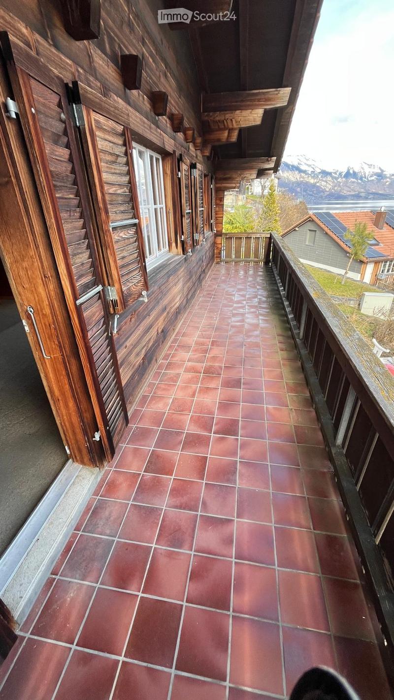 Balcony with red tiled flooring, wooden railings, and wooden walls, overlooking a scenic view of mountains and a lake.