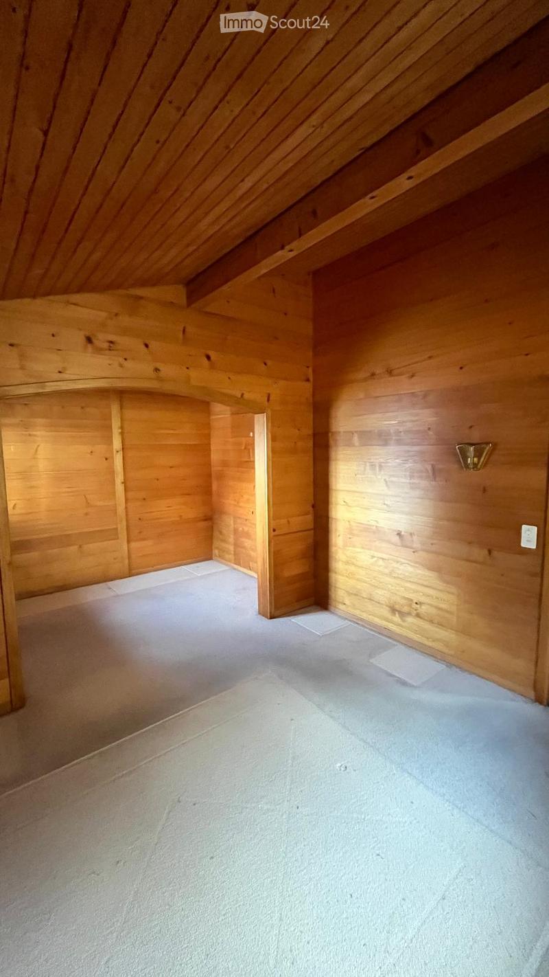 Empty attic space with wooden walls and slanted wooden ceiling, carpeted floor, and a small doorway leading to another area.