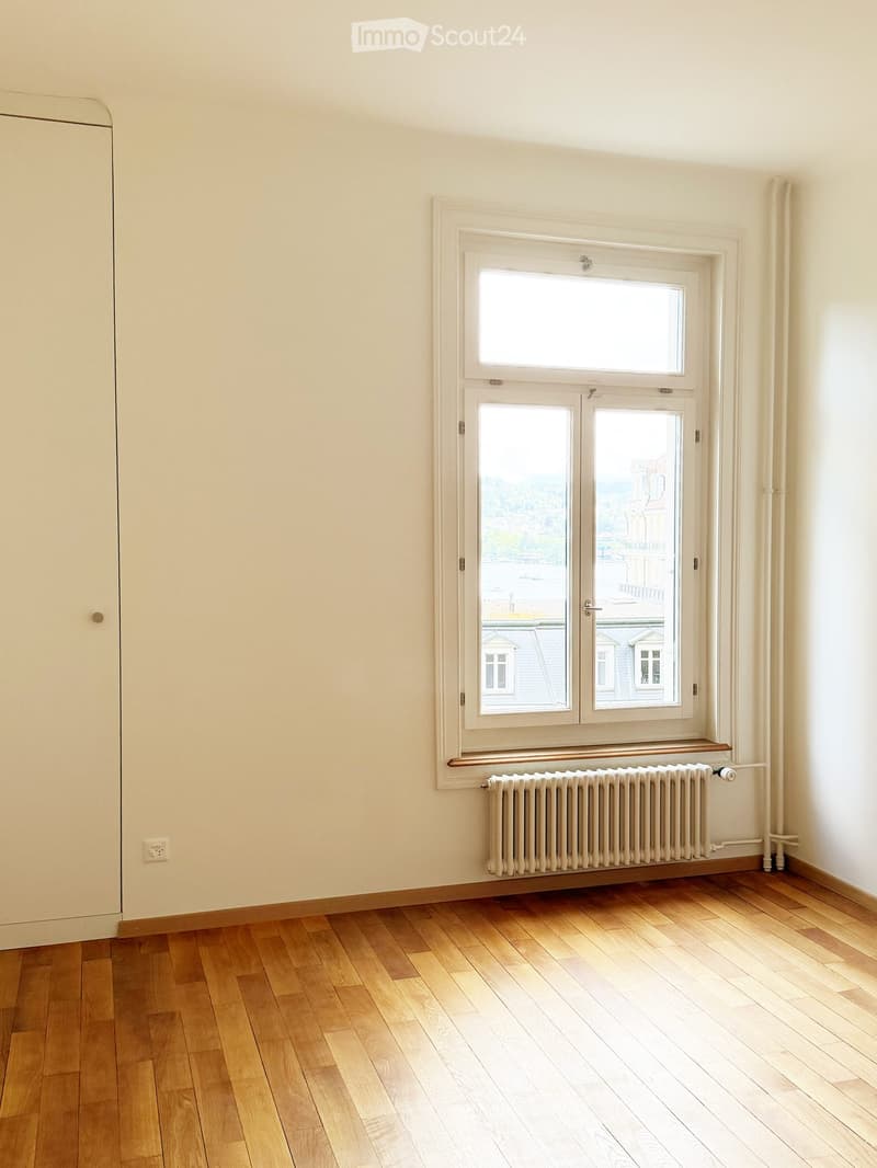 Empty room with wooden flooring, white walls, a large window with double doors, and a radiator below the window.