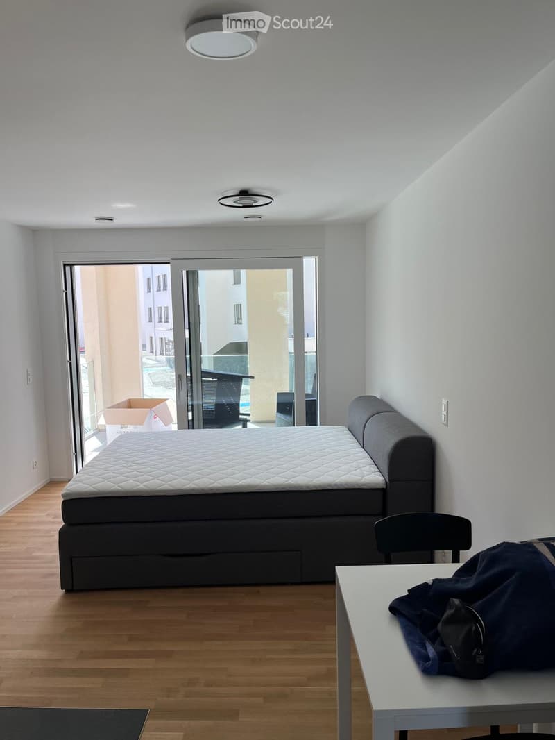 A modern, minimalist bedroom with a large bed, a chair, a small table with clothing, and a sliding glass door leading to a balcony.