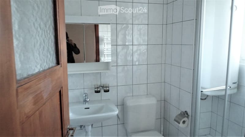 Bathroom with white tiles, a sink, a toilet, a mirror, and a toilet paper holder. The door is wooden with a glass panel.