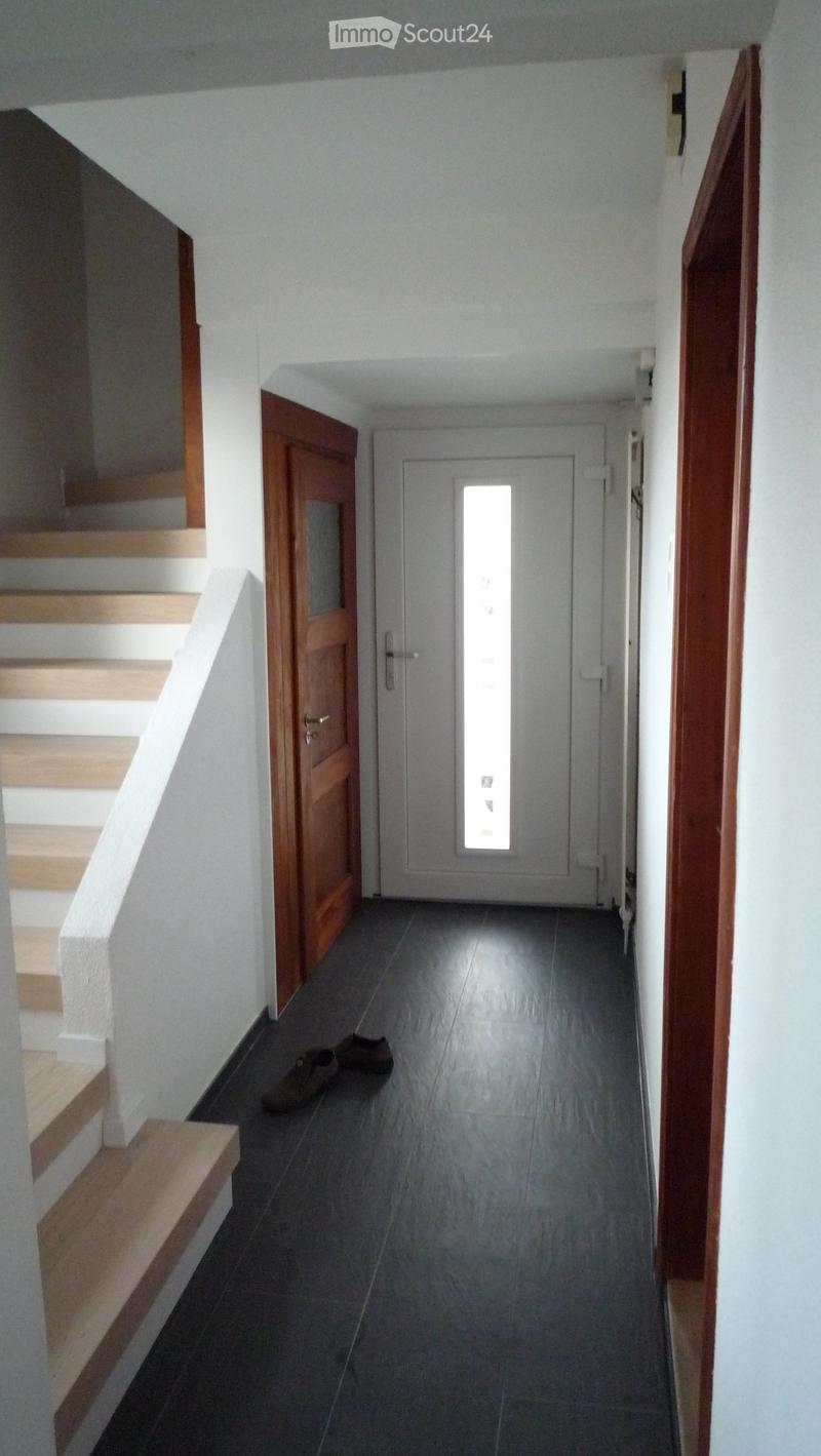 A narrow hallway with a wooden staircase on the left and a closed door on the right, featuring dark tiled flooring and white walls.