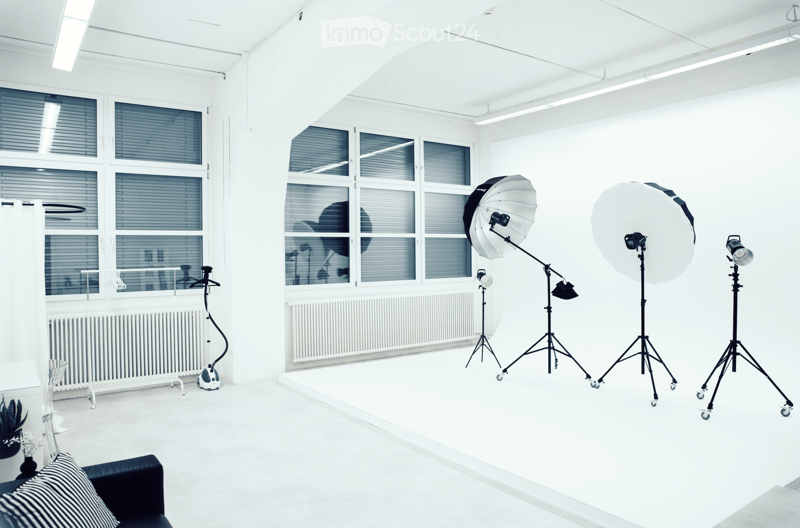 Bright studio with large windows, white walls, and a sofa with a striped pillow. Photography equipment including tripods, umbrella lights, and a camera is set up.