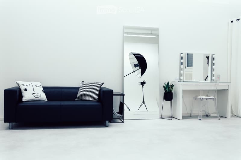A modern, minimalist room with a dark blue sofa, a large mirror, a vanity with a chair, a small table, and a potted plant. A camera on a tripod is positioned near the mirror.