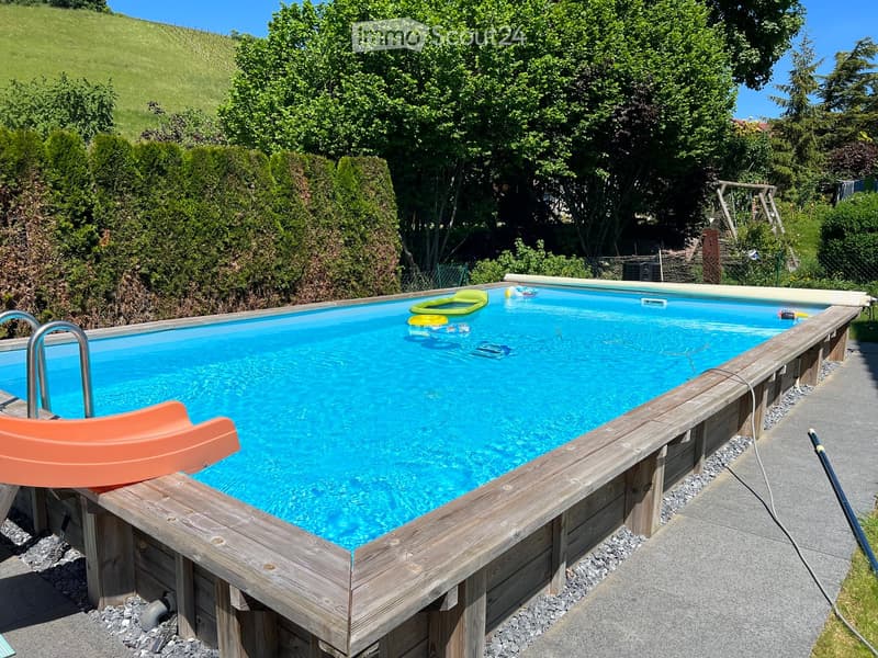 A rectangular swimming pool with clear blue water, surrounded by wooden decking and greenery. The pool features a slide on one side and inflatable toys floating in the water.