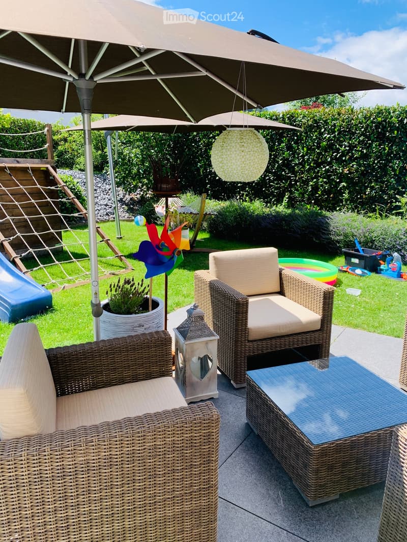 A sunny outdoor patio with wicker furniture, including a sofa, chairs, and a coffee table, under a large umbrella. The area features a pinwheel, a lantern, a potted plant, and a view of a grassy yard with a slide and toys.