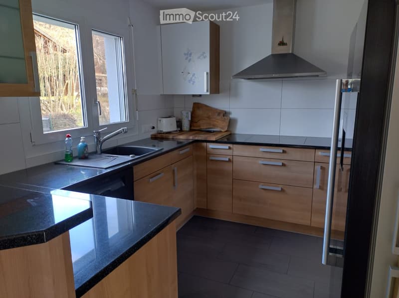 Modern kitchen with light wooden cabinets, granite countertops, stainless steel sink, and a built-in range hood. Features include a window with a view of greenery, a toaster, and a wooden cutting board on the counter.