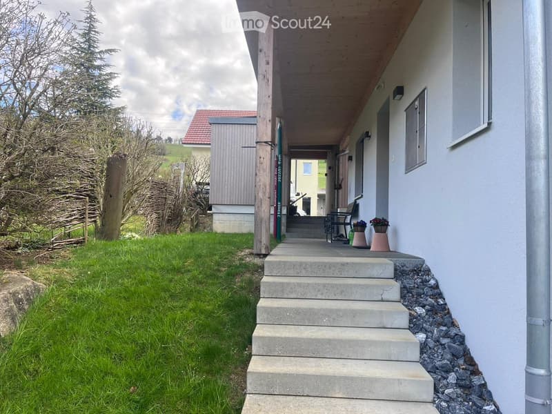 A grassy lawn leads to a concrete staircase that ascends to a wooden porch with a bench and potted plants. The house has a white exterior with a red-tiled roof, and the surrounding area includes trees and a neighboring building.