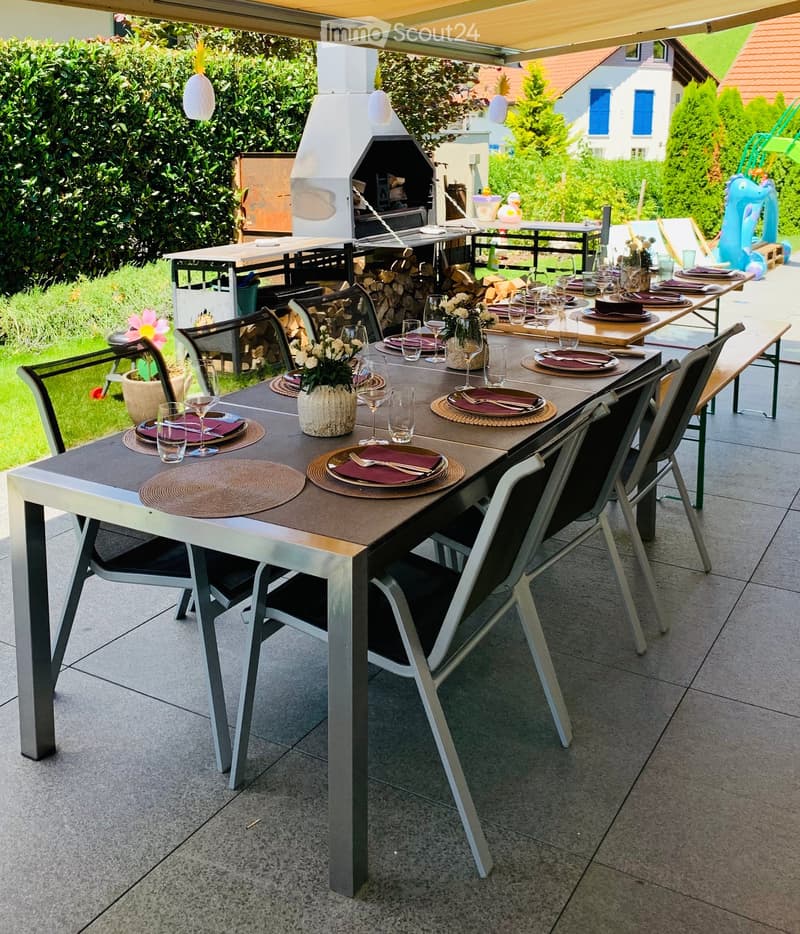 A well-maintained outdoor dining area with a long rectangular table set for a meal, surrounded by chairs, a barbecue, and a neatly arranged garden.