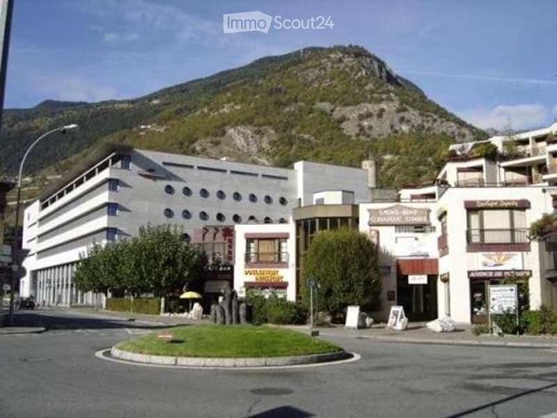 Modern multi-story building with a rounded facade, circular driveway, and surrounding greenery, set against a mountainous backdrop.