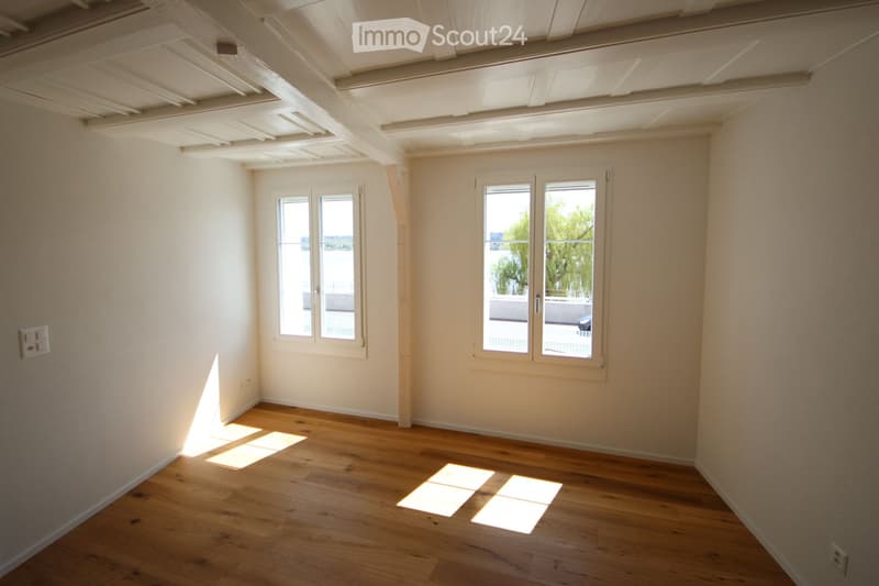 Empty room with wooden flooring, white walls, and large windows allowing natural light to enter. The room features exposed wooden beams on the ceiling and a visible electrical outlet on the wall.