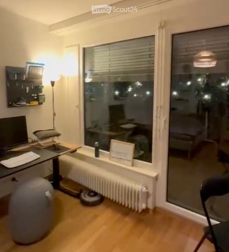 A modern, well-lit room with wooden flooring, large windows, a desk with a computer setup, a radiator, and a picture frame on the windowsill.