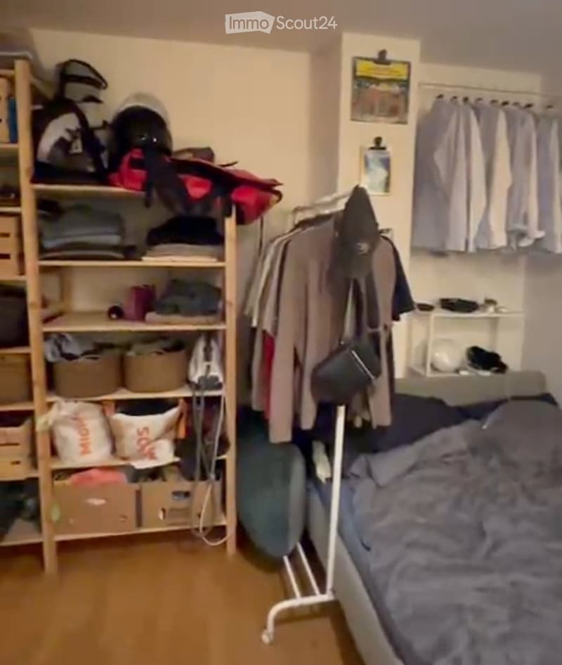 A small, furnished bedroom with a bed, a clothing rack, a shelf filled with various items, and a wardrobe. The room features wooden flooring and has a cozy, lived-in feel.