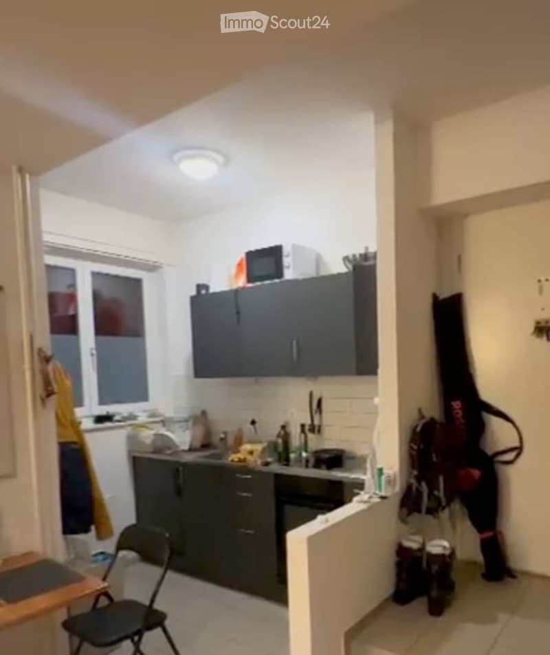 Modern kitchen with dark grey cabinets, a microwave, a sink, and a small table with a chair. The space is well-lit with white walls and a window providing natural light.