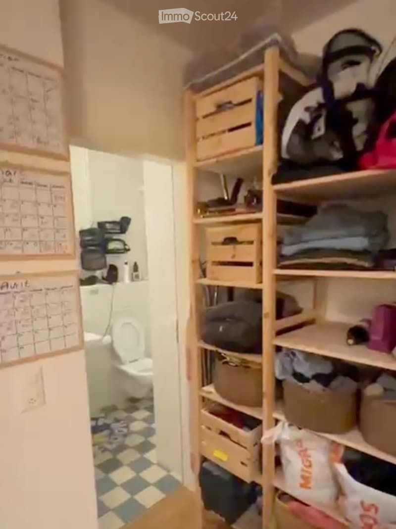 A small bathroom with a toilet, checkered floor, and a wooden shelf unit filled with storage boxes and folded clothes. Calendars are visible on the wall.