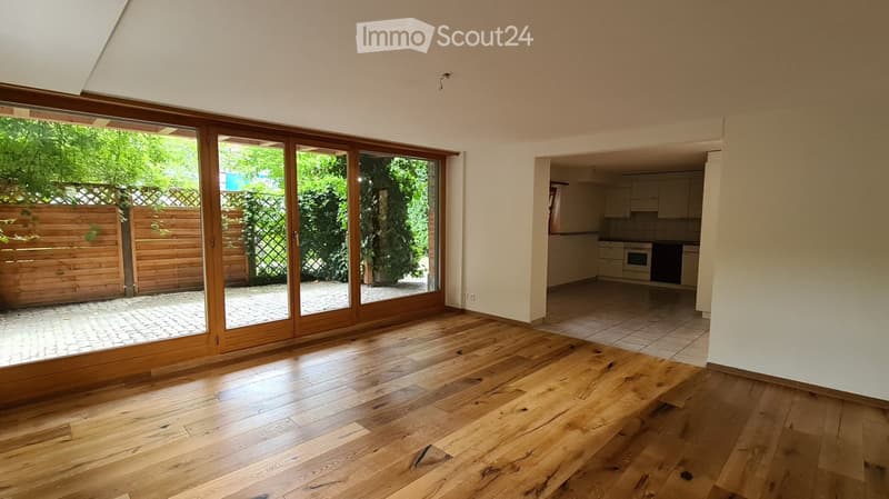Bright, unfurnished room with wooden flooring, large sliding glass doors leading to an outdoor area, and an open doorway to a kitchen area.