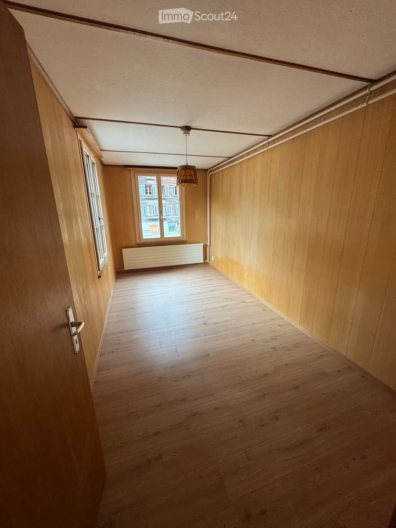Empty hallway with wooden flooring, wooden walls, and a hanging light fixture. There is a window at the end showing an exterior view.