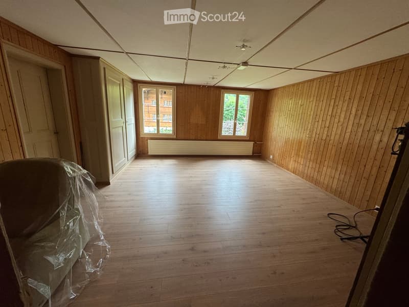 Empty room with wooden walls and floor, two windows, and a radiator. No furniture or equipment present.