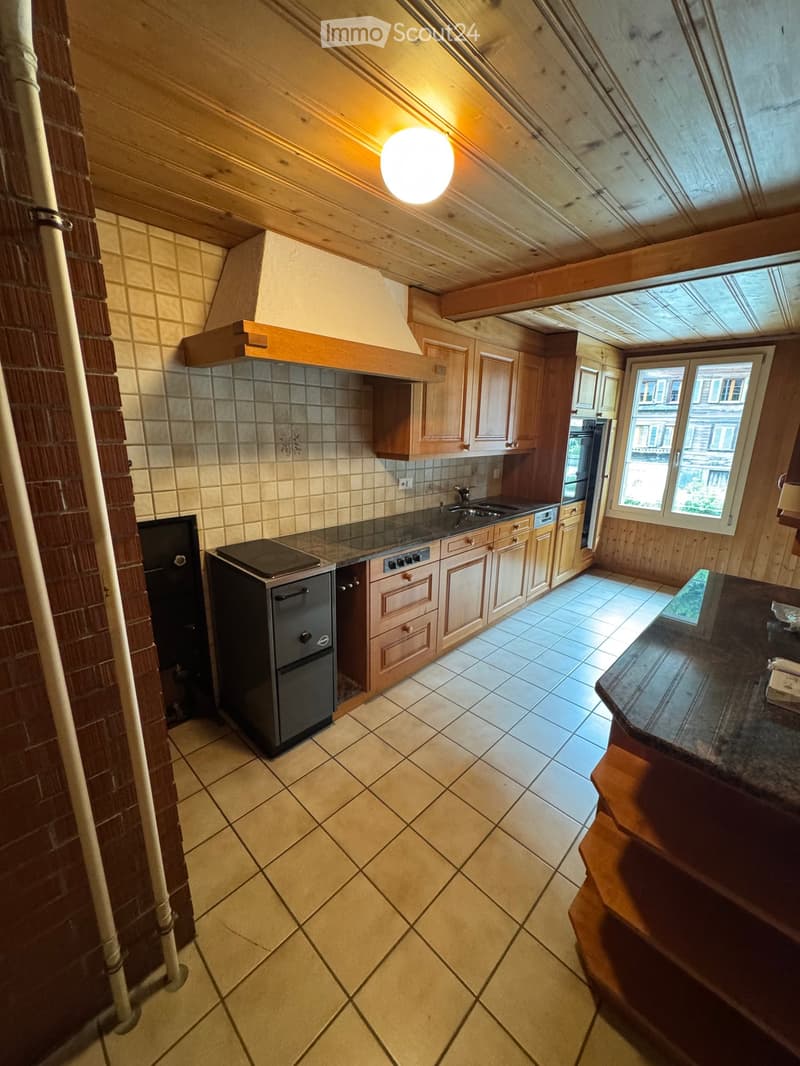 Kitchen with wooden cabinets, tiled floor, and appliances including a refrigerator, oven, and sink. Natural light from a window on the right.