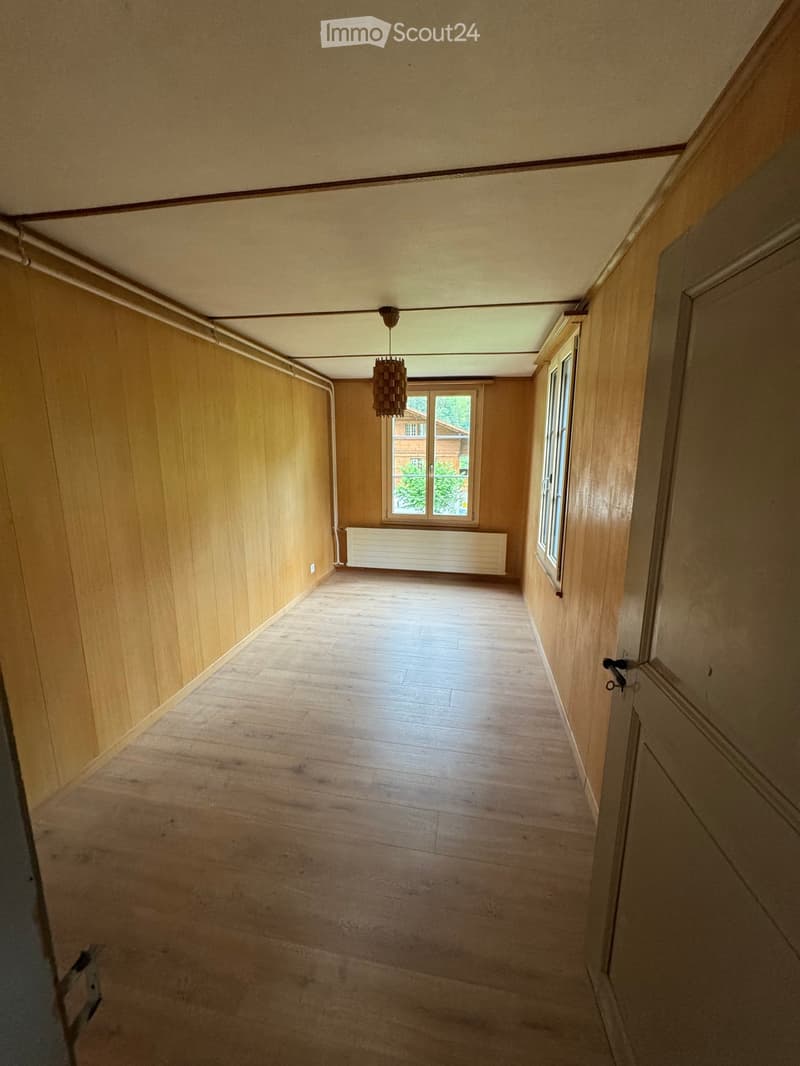Empty hallway with wooden walls and flooring, a single window at the end, and a hanging light fixture.