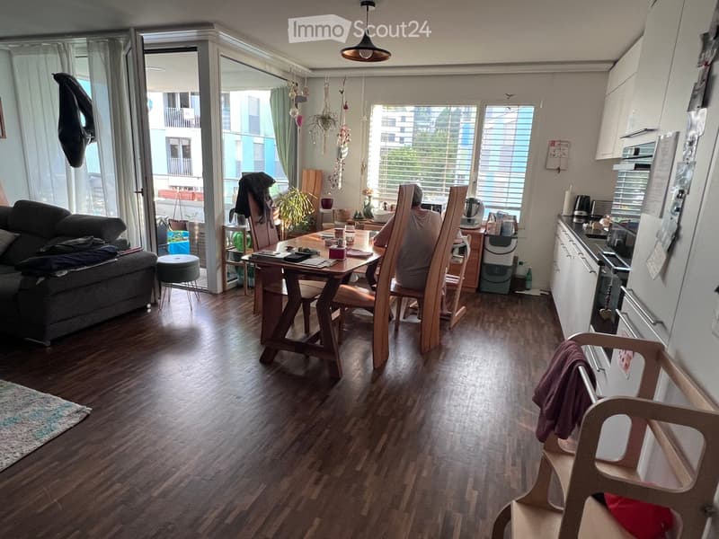 Open-plan living and dining area with wooden flooring, a sofa, dining table with chairs, kitchen area with cabinets and appliances, and large windows with a view of the balcony.