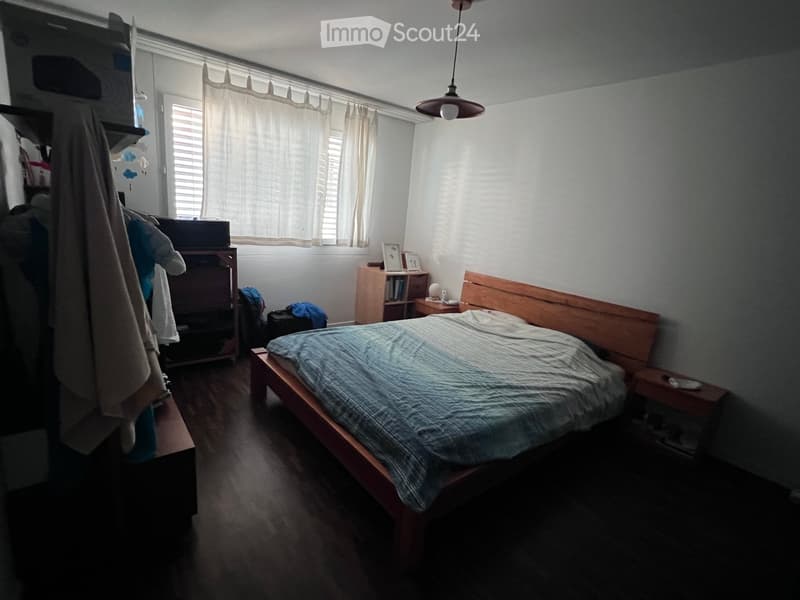 Bedroom with a wooden bed, nightstand, bookshelf, and wardrobe. The room has wooden flooring and a window with curtains.
