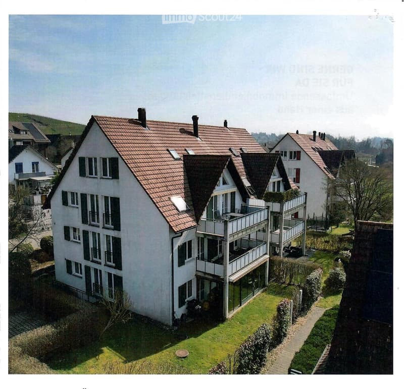 A two-story house with a red-tiled roof, multiple windows, and a balcony. The house is surrounded by a well-maintained lawn and garden, with neighboring houses visible in the background.