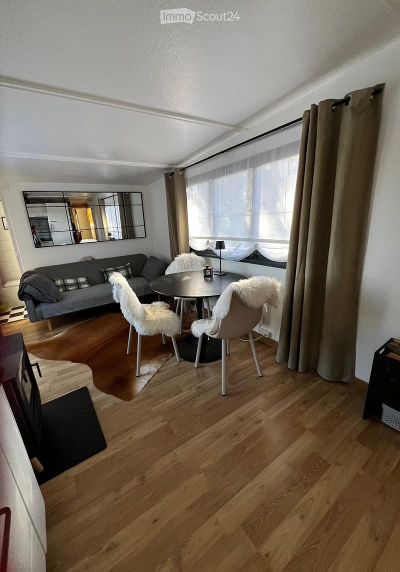 Modern living room with wooden flooring, a grey sofa, a round table with two chairs, a mirror on the wall, and a lamp near the window.
