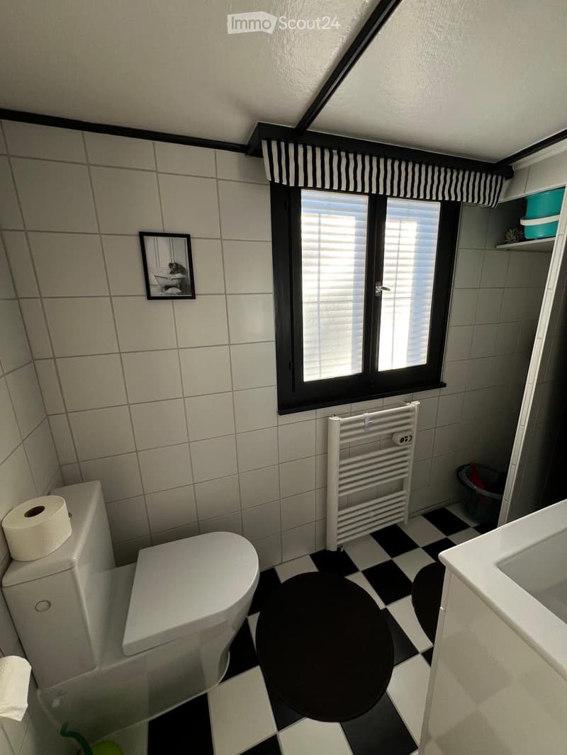 Bathroom with a toilet, sink, radiator, and a window with blinds. The floor has a black and white checkered pattern, and there is a mirror on the wall.