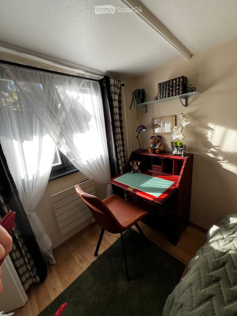 Cozy bedroom with wooden floor, large window with white curtains, desk with chair, lamp, mirror, bookshelf, plant, teddy bear, flower vase, and clock.