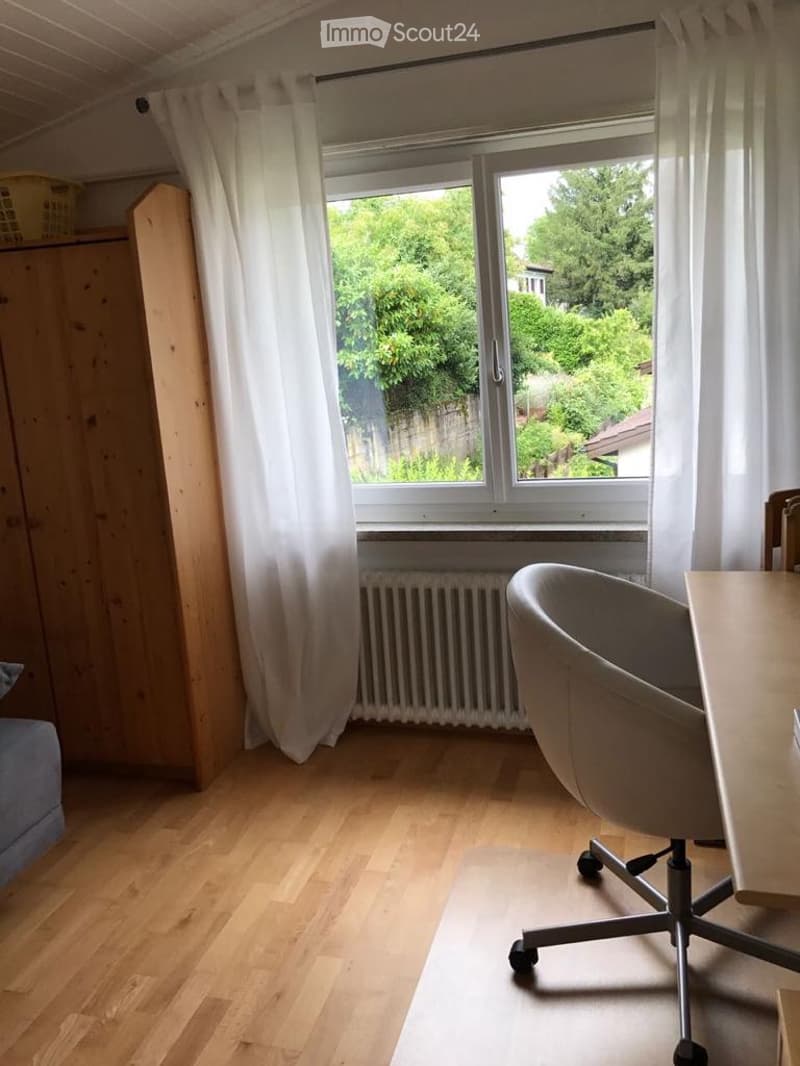 Bright room with wooden flooring, large window with white curtains, a grey office chair, a wooden table, and a radiator below the window.
