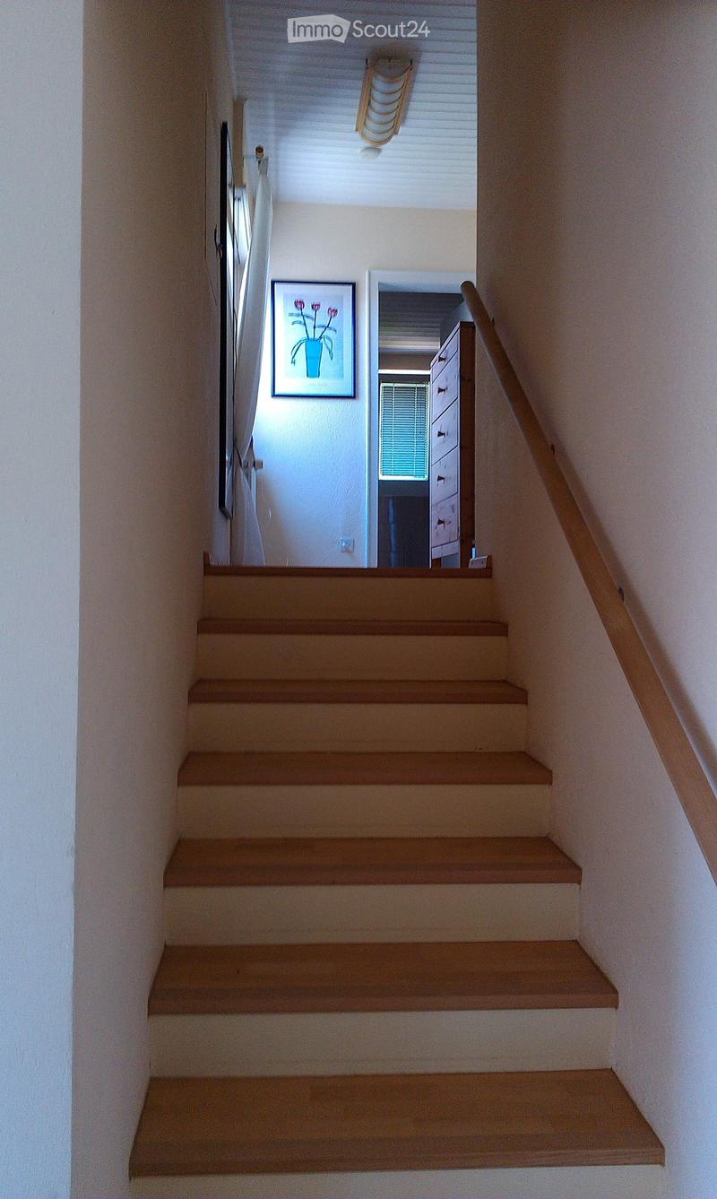 Wooden staircase with a handrail, light-colored walls, and a framed picture on the wall. The staircase leads to an open area with a window and a glimpse of a room with a dresser.
