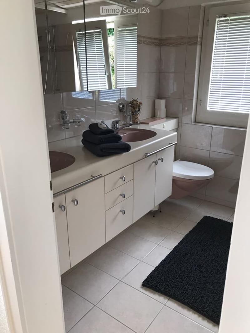Modern bathroom with double sink, large mirror, white cabinets, and a toilet. Features a black rug on the floor and a window with blinds.
