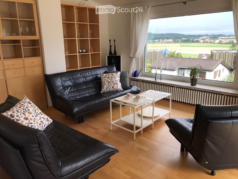 Modern living room with large windows offering a scenic view, featuring a black leather sofa, armchair, white coffee table, and wooden shelving unit.