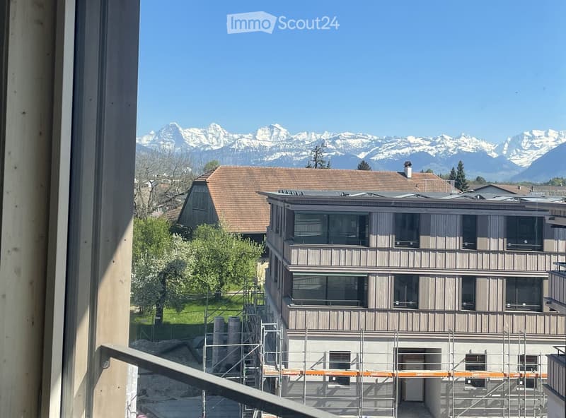 Modern multi-story building with balconies and large windows, surrounded by greenery and snow-capped mountains in the background.