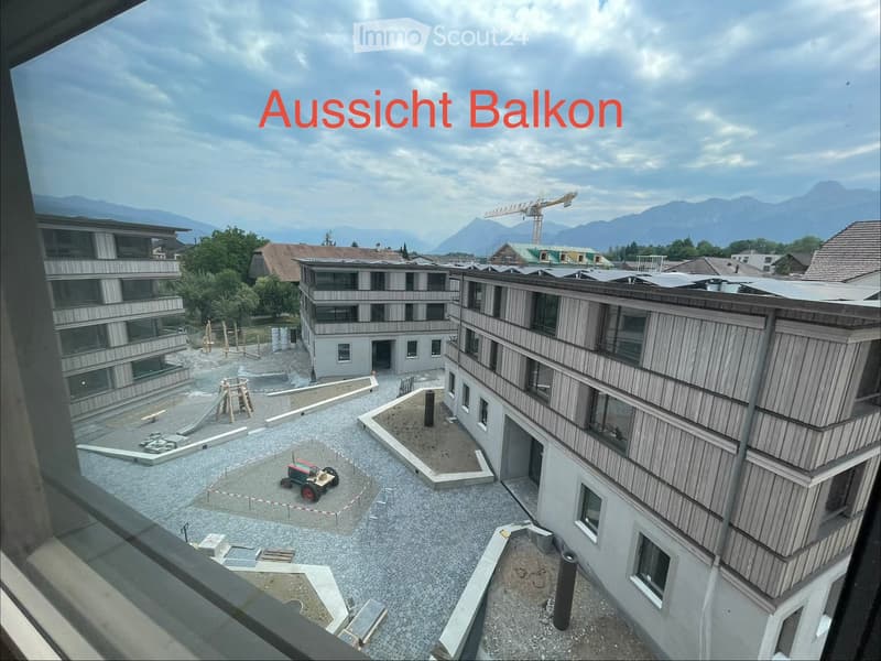 View from a balcony showing a modern building complex with wooden facades, a playground area, and distant mountains under a partly cloudy sky.