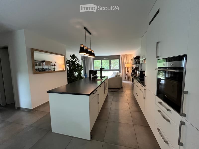Modern kitchen with white cabinets, black countertop, integrated appliances, and a living area visible through an open doorway.