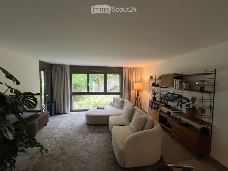 Modern living room with a large window, cream-colored sofa, ottoman, wooden shelf, and decorative plants.