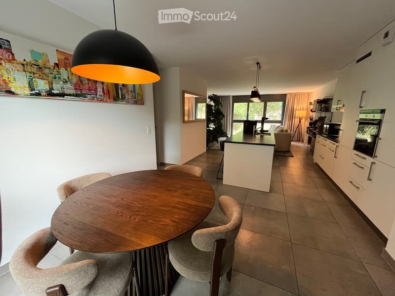 Open-plan living and dining area with a round wooden dining table, four upholstered chairs, a modern kitchen island, white cabinets, a sofa, and a large pendant light fixture.