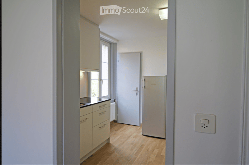 The image shows a modern, minimalist kitchen with light-colored cabinets, a stainless steel refrigerator, and a sink. The floor is made of light wooden planks, and the space is well-lit with natural light coming through a window.