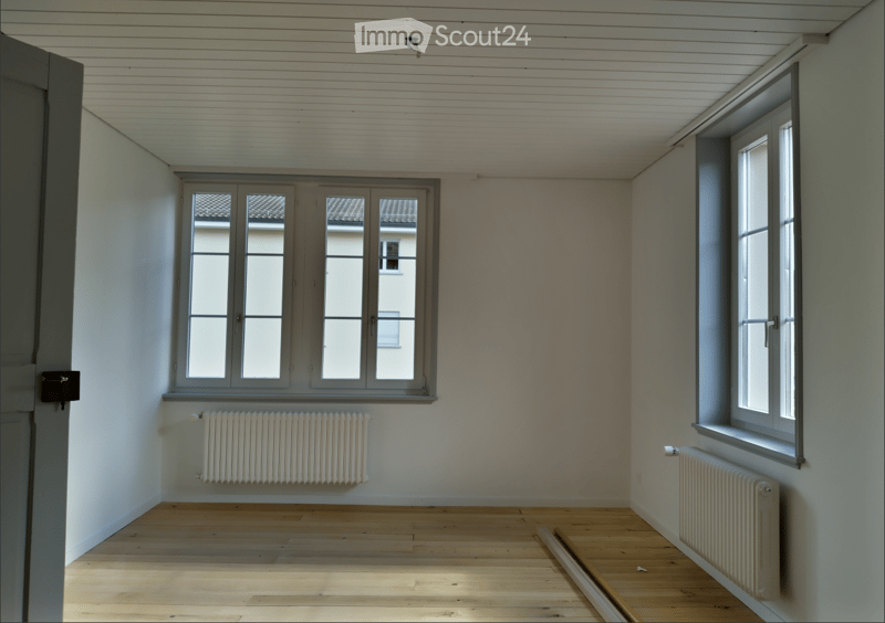 Empty room with wooden flooring, white walls, and large windows. Radiators are present on both sides of the room.
